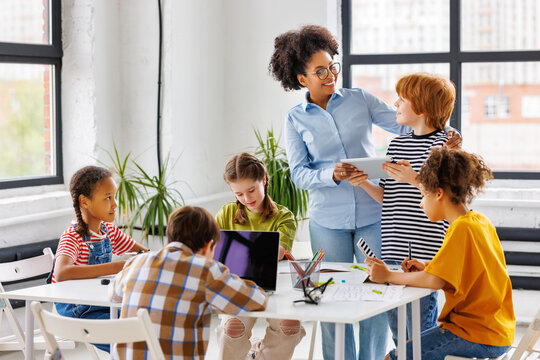African american teacher smiling and checks work of student looking at tablet during lesson.