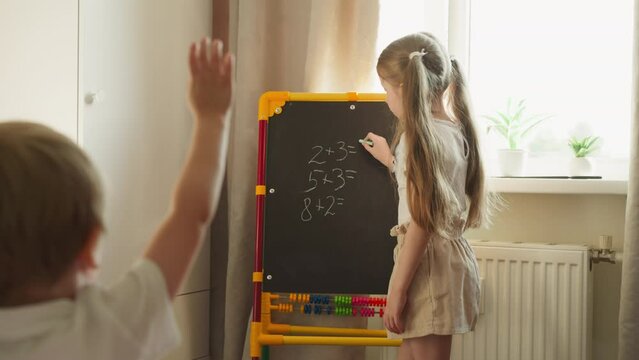 Little schoolgirl stands at blackboard in room teaching toddler brother to solve math problems slow motion. Boy thinks and raises hand telling answer