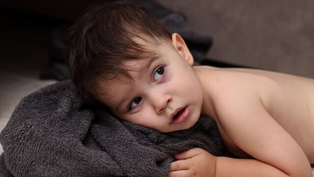 adorable baby boy kid lying on sofa with coverlet,gray tones.child kissing sole toe, leg up.toddler making funny faces sitting at home on bed, napping time 4k