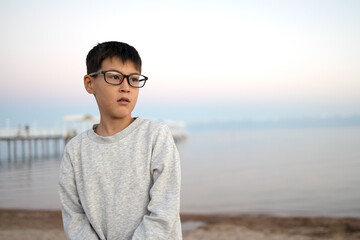 Attractive male teenager dressed in trendy sweater and wearing glasses standing on the beach at...