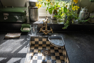 traditional old style country house kitchen area with chequered sink basin, brass tap faucet and accessories lit naturally from the window. Soap in a dish alongside a hand cloth and flowers
