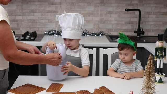 siblings boys doing with granny gingerbread christmas house.preschooler child wearing toque chef  hat,apron,mixing cream,licking from wires beaters.toddler with tree headband,modern kitchen 4k