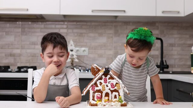 siblings boys doing with granny gingerbread christmas house.preschooler child wearing toque chef  hat,apron,mixing cream,licking from wires beaters.toddler with tree headband,modern kitchen 4k