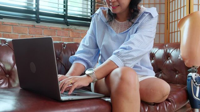 Young asian woman disabled sitting on sofa using laptop computer with prosthetic leg in living room at home, female working on notebook and prosthesis with optimistic, lifestyles and disability.