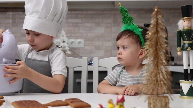 siblings boys doing with granny gingerbread christmas house.preschooler child wearing toque chef  hat,apron,mixing cream,licking from wires beaters.toddler with tree headband,modern kitchen 4k