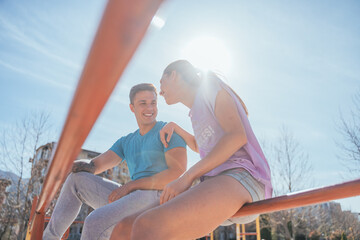 Lovely sports couple taking a break from workout while sitting on a horizontal metal bar. They are having fun conversation and smiling