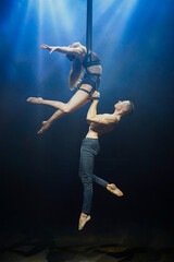 An aerial straps duo performs dangerous tricks mid-air, illuminated by strong blue and white lights against a dark background, clad in minimal athletic attire, showcasing a blend of power and grace