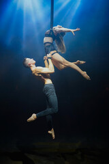 An aerial straps duo performs dangerous tricks mid-air, illuminated by strong blue and white lights against a dark background, clad in minimal athletic attire, showcasing a blend of power and grace
