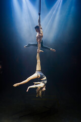 Aerial straps duo performance: a man and woman execute graceful acrobatic feats in mid-air against a black background, dressed in black and illuminated by a white-blue glow