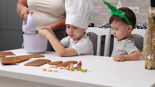 siblings boys doing with granny gingerbread christmas house.preschooler child wearing toque chef  hat,apron,mixing cream,licking from wires beaters.toddler with tree headband,modern kitchen 4k
