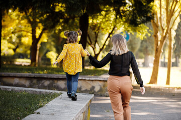 Obraz premium Mothers day. Happy young mom is holding by hand and walking with adorable little daughter at autumn park. Mom with kid are having fun, playing at weekend. Outdoors vacation activity. Children's day.