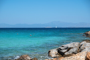 Greece Agistri Island. Rocky beach, people swimming sailboat sails in sea water, blue sky sunny day