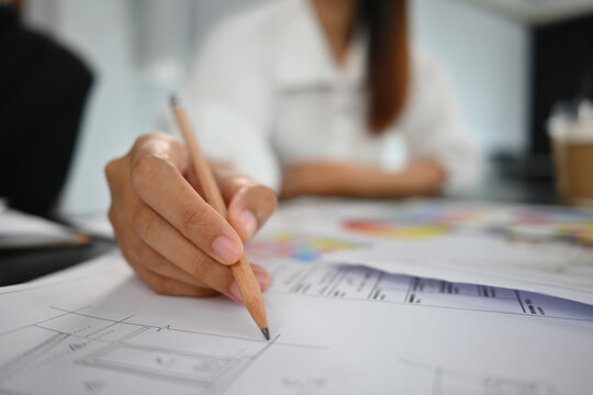Close-up image of Young interior designer or architect holding a pencil and pointing at blueprint placed on meeting desk, Creative team collaborative planning.
