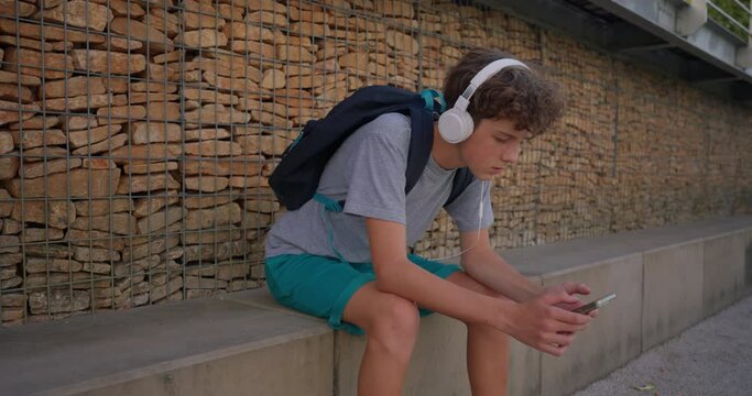 A cute teenager sits in a park on a bench and listens to music in headphones while looking at the phone