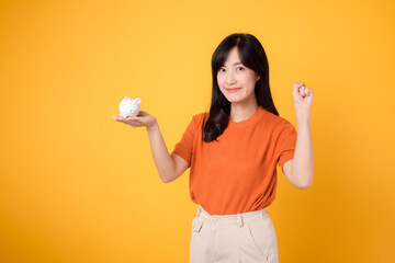 Joyful Asian woman in her 30s holding piggy bank, showing zero hand sign, on vibrant yellow background. Wealth saving concept.