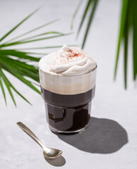 Iced coffee in a glass with foam on a light background with palm leaves, spoon and morning shadows.