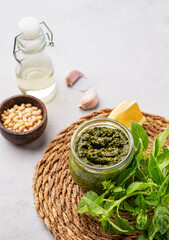 Traditional Italian pesto in a jar with green basil, pine nuts, cheese and olive oil on a light background. A classic sauce for spaghetti or bruschetta.