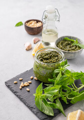 Traditional Italian pesto in a jar with green basil, pine nuts, cheese and olive oil on a slate board on a light background. A classic sauce for spaghetti or bruschetta.