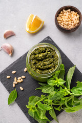 Traditional Italian pesto in a jar with green basil, pine nuts, cheese and olive oil on a slate board on a light background. A classic sauce for spaghetti or bruschetta. 