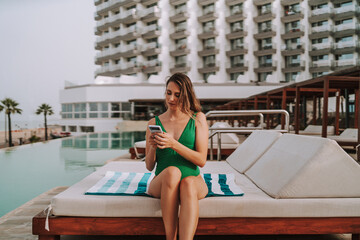 Chica joven en hamaca con smartphone tomando fotografías y leyendo en piscina de hotel de lujo