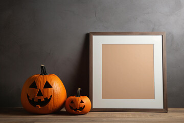 An empty square frame for mockup stands on the table near the jack o lantern pumpkin. Grey wall background. Halloween decor.