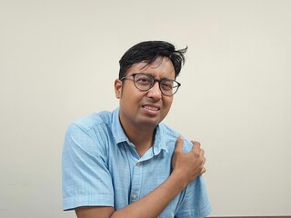 Indian man with glasses in a blue shirt showing shoulder pain against a gray background