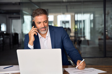 Middle aged Latin or Indian businessman having call on smartphone with business partners or clients. Smiling mature Hispanic man sitting at table talking by mobile cellphone and make notes in office.