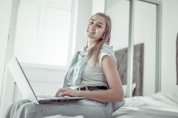 Young smiling woman using laptop while sitting on bed in bright room