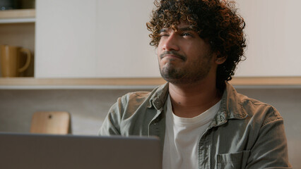 Pensive ethnic guy with curly hair thinking idea typing laptop at kitchen. Thoughtful Indian man...