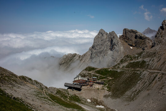 The mountain station of the Karwendelbahn is nestled among rugged peaks and alpine meadows on the Mittenwald High Trail, surrounded by drifting clouds under a clear blue sky.