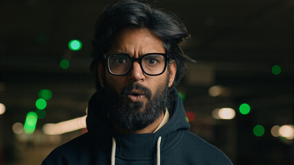 Close up Indian shocked bearded guy in glasses astonished looking at camera at dark underground...