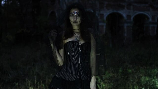 A mysterious Gothic woman in a black dress with a corset, her head is covered with a black veil. Model posing in front of an old abandoned building. Autumn forest. Halloween concept. Horizontal video