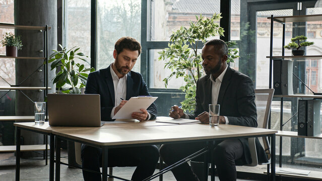 Moving camera diverse partners in office sign contract handshake cooperation Multiethnic Caucasian and African American business people men sit at desk signing document shake hands success partnership