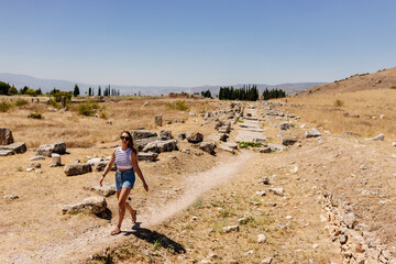 A young woman in a striped top and blue denim shorts walks along a picturesque road between the ruins of an ancient city. Journey to the old city. Hierapolis, Pamukkale, Türkiye - July 29, 2023