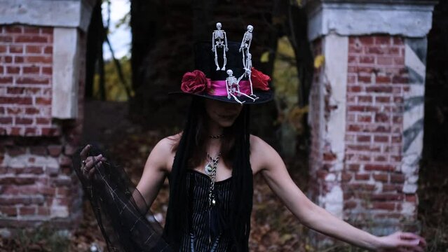 Female Halloween look. A woman in a black dress with a corset, a top hat decorated with skeleton figures poses for the camera against the backdrop of an old abandoned building. Horizontal slow motion 