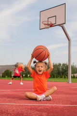 A cute funny little laughing kid is sitting at the stadium holding a basketball over her head. Children play ball on the playground