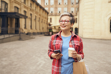 Smiling mature woman walking with her shopping bags, drinking coffee and use phone in city street 