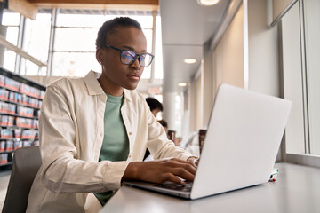 Busy short-haired African girl student using laptop technology sitting in university campus library. Serious young Black woman elearning looking at computer advertising hybrid work and elearning.