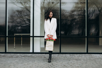 Attractive brunette woman walking glass office building in moody cloudy windy weather with bouquet of red tulips flowers. International Women`s Day 8th March concept
