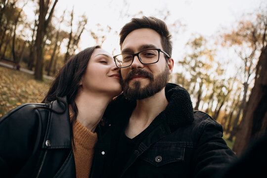 Heterosexual caucasian young loving couple walking outside make selfie in sunny weather, hugging smiling kissing laughing spending time together. Autumn, fall season, orange yellow red maple leaves


