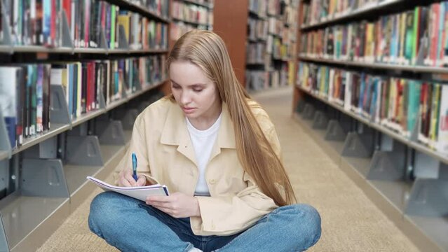 European girl student sitting on floor among bookshelves in university campus library learning, schoolgirl doing college course work study, doing research, writing notes reading book.