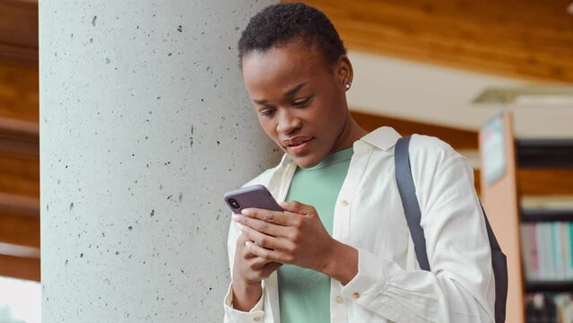 African Black teen girl college generation z student with backpack looking at smartphone using mobile cell phone modern tech device standing in university campus with cellphone.