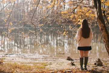 A young woman with long hair in a beige sweater, a short black skirt and rubber boots is standing...