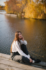 Fototapeta na wymiar A woman with long hair in a white shirt and grey trousers is sitting on a wooden masonry near the lake in autumn. 