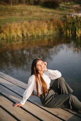 A woman with long hair in a white shirt and grey trousers is sitting on a wooden masonry near the lake in autumn. 