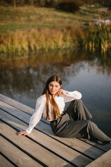 A woman with long hair in a white shirt and grey trousers is sitting on a wooden masonry near the lake in autumn. 