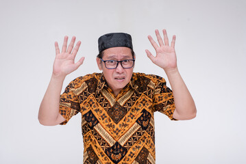 A fearful southeast asian muslim man puts his hands in the air and surrenders. Wearing a batik shirt and songkok skull cap with hand on chest. Isolated on a white background.