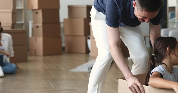 Happy parents playing with kid among moving packages in new apartment, Joyful dad pushing forward cardboard box with cheerful little girl with open arms inside, enjoying relocation funny activity