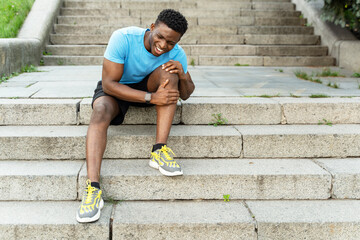 Sad african american man in blue t shirt and sneakers holding knee pain in leg sitting on stairs