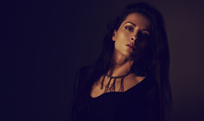Fototapeta premium Beautiful woman in black dress posing in fashion necklace on the neck. Long hairstyle on dark shadow background. Toned portrait. Closeup.